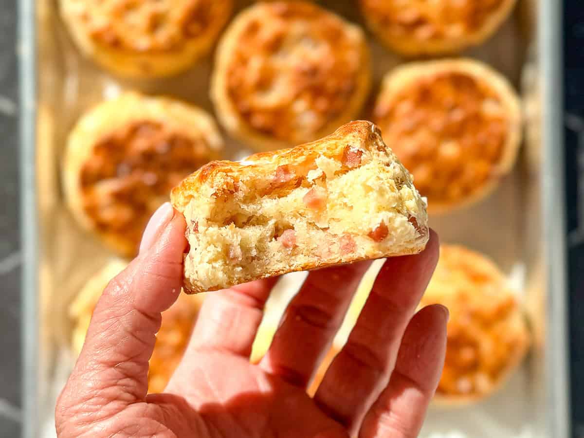 Hand holding a ham and cheese biscuit with a bite taken out, showing the fluffy and flaky interior.