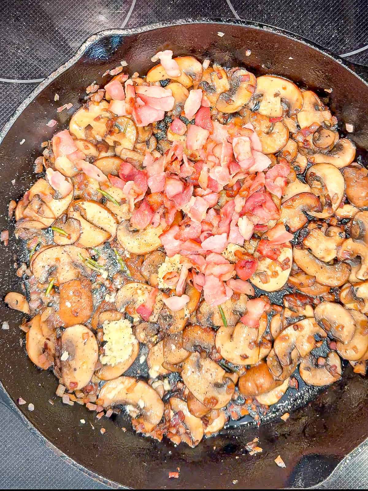 Mushrooms bacon onions and garlic in a black pan.