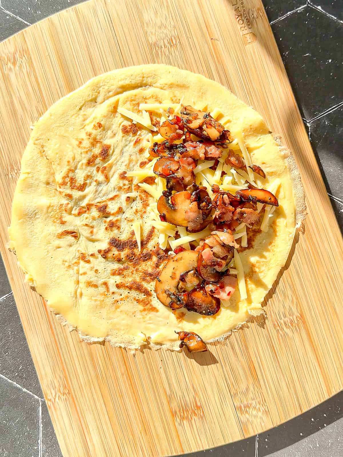 A crêpe with shredded cheese and a mushroom mixture on a wooden cutting board.