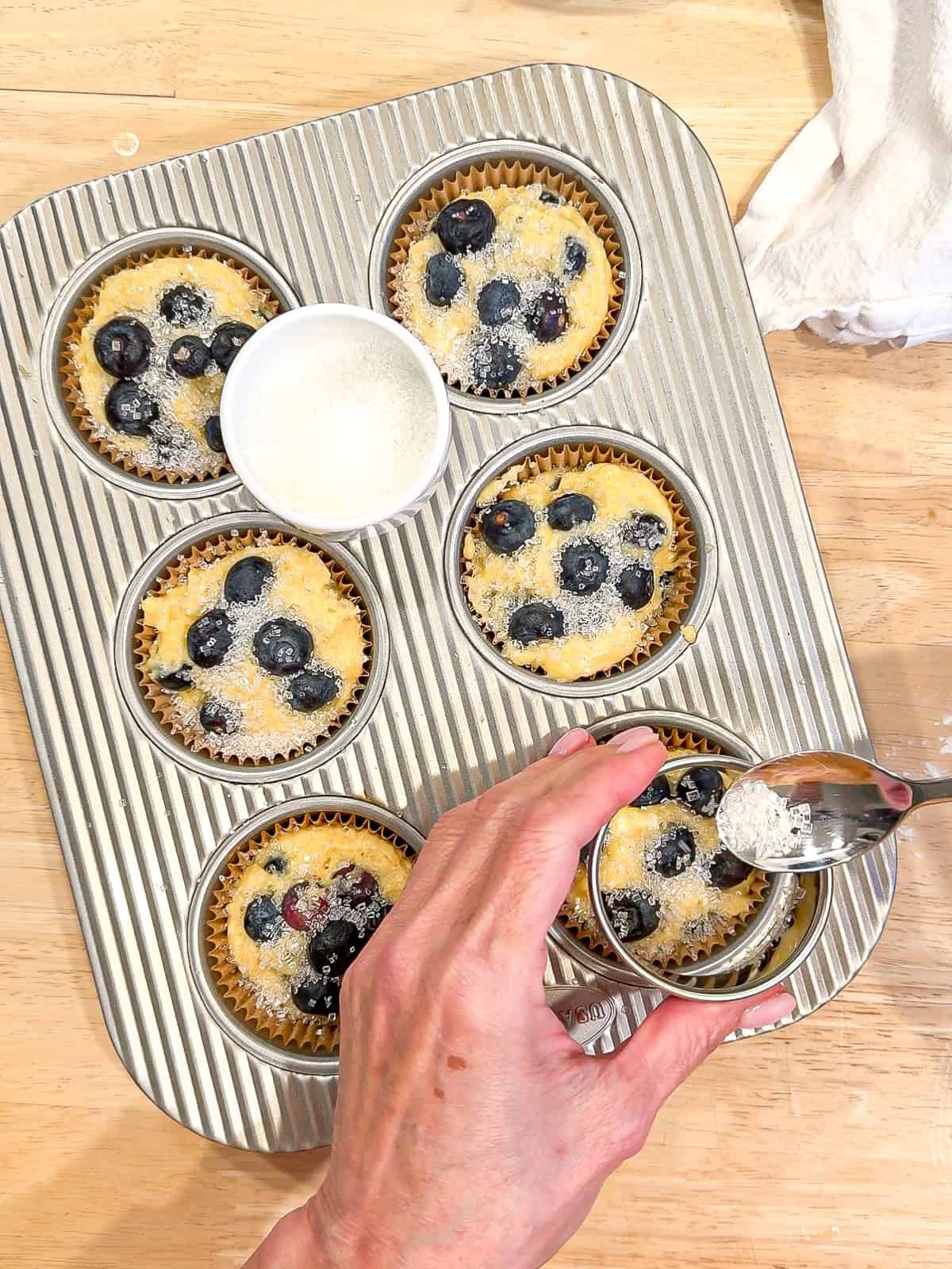 Sprinkling coarse sugar on top of muffin batter in a muffin pan.