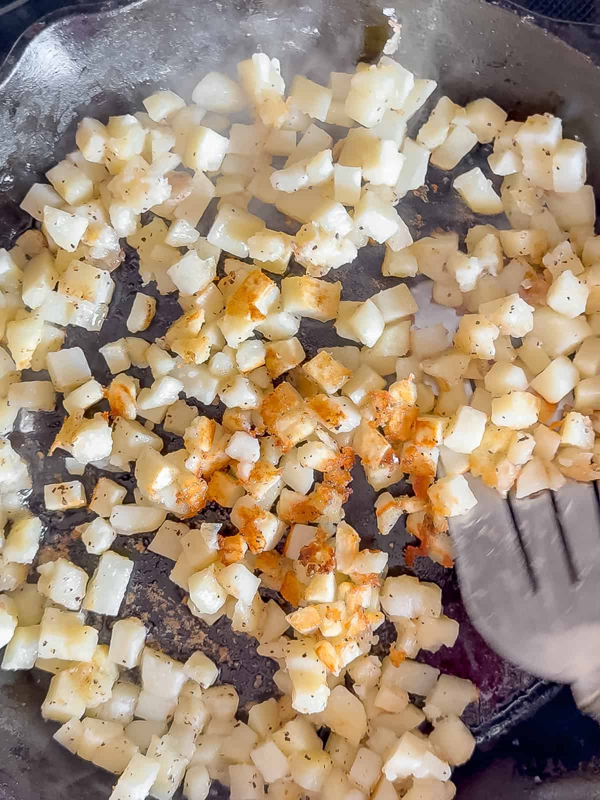 Flipping over crispy potatoes in a cast-iron pan using a silver spatula.
