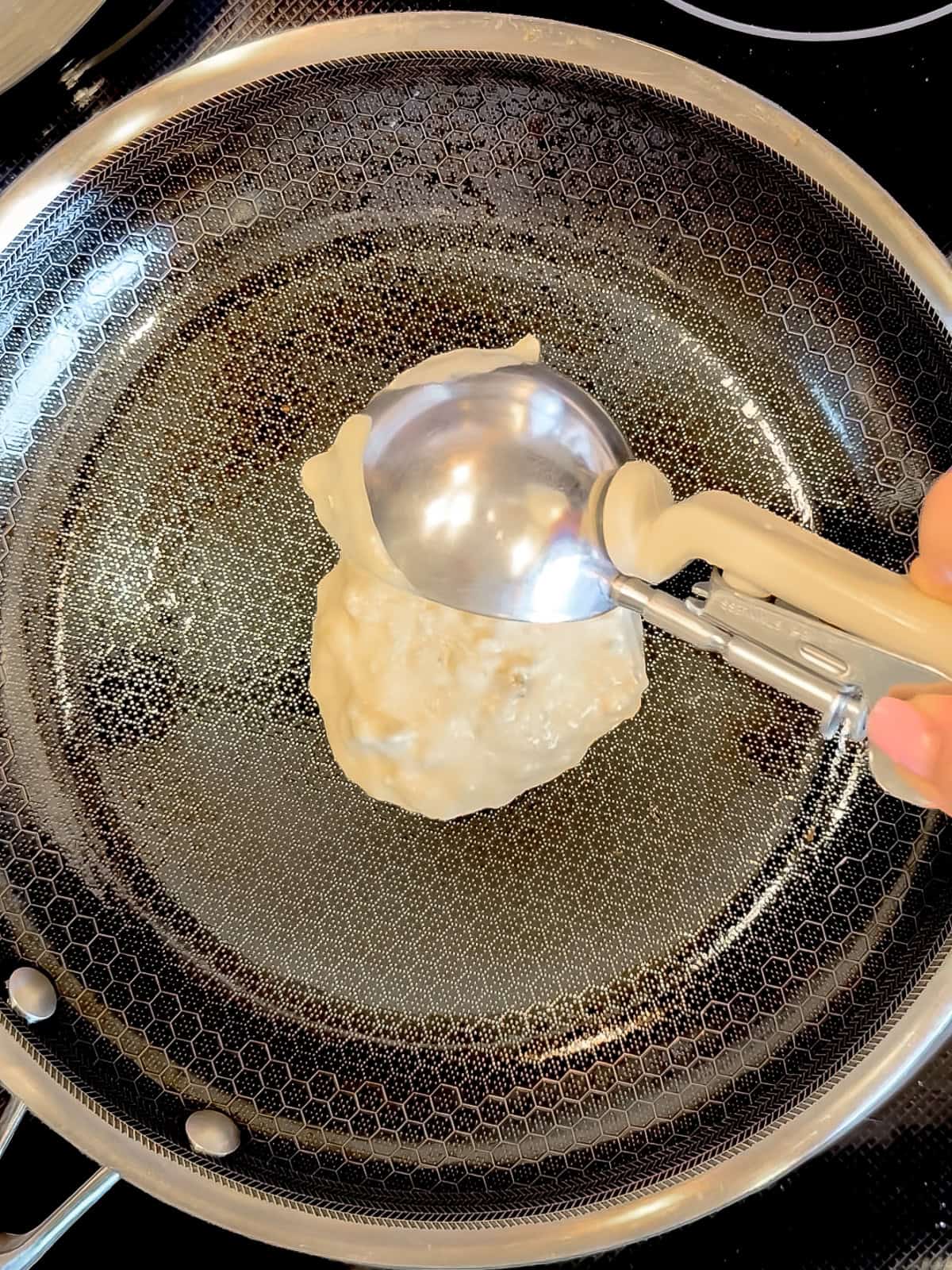 Using an ice cream scoop to portion pancake batter into a black pan.