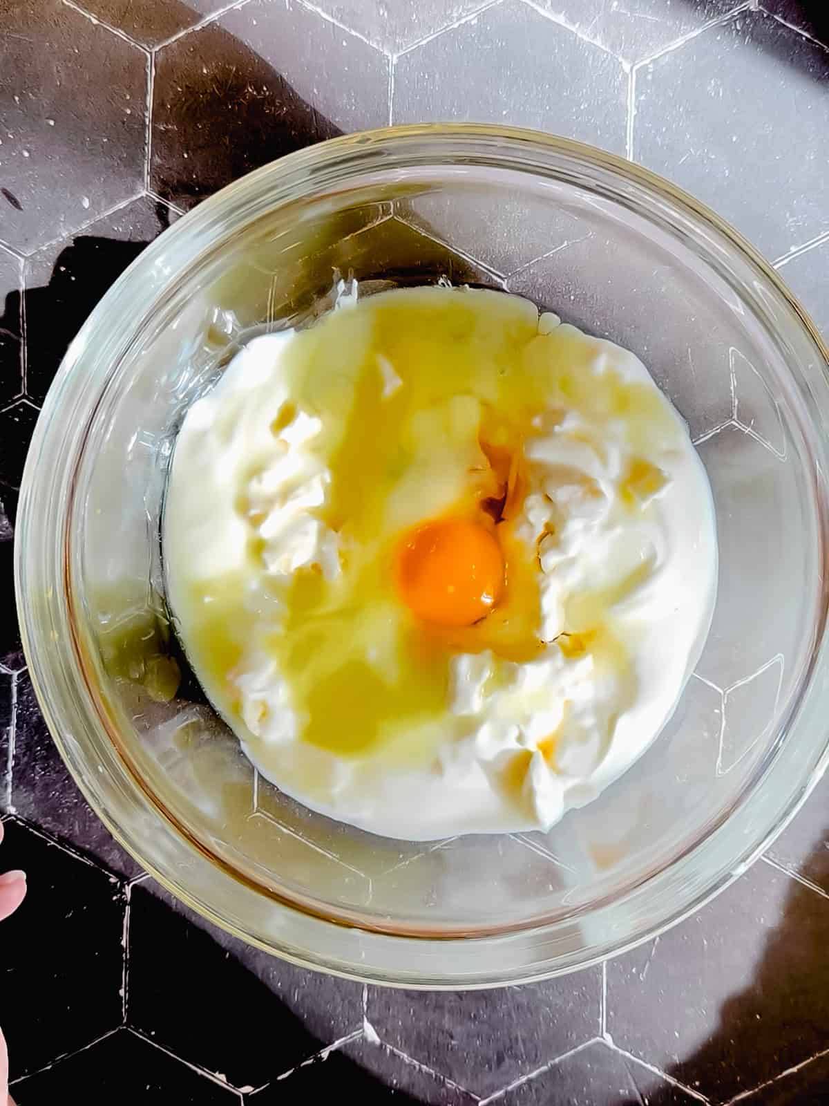 Adding yogurt, maple syrup, vanilla, an egg, and a melted butter to a mashed banana in a glass bowl.