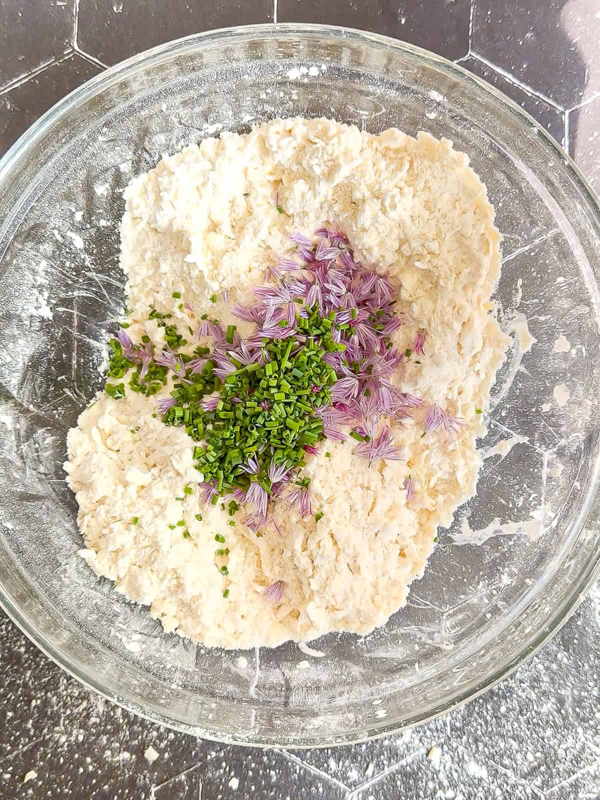 Adding chive blossoms and chives to dry ingredients in a glass bowl.