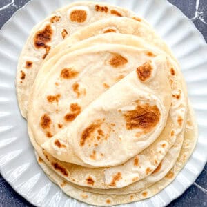 A stack of flour tortillas on a white scalloped edge plate placed on a black tile background.