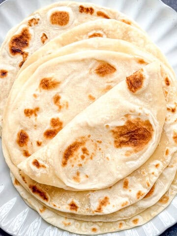 A stack of flour tortillas on a white scalloped edge plate placed on a black tile background.