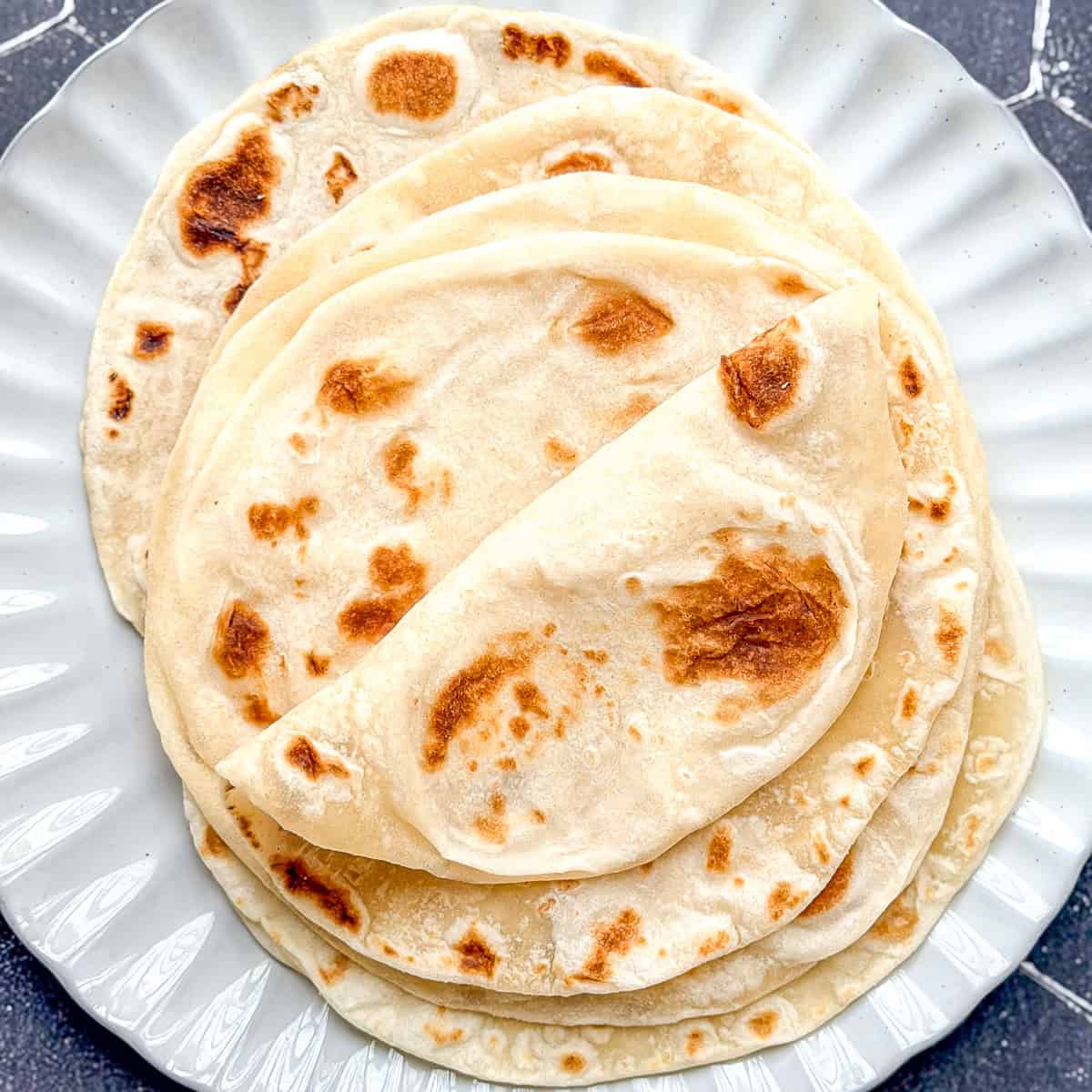 A stack of flour tortillas on a white scalloped edge plate placed on a black tile background.