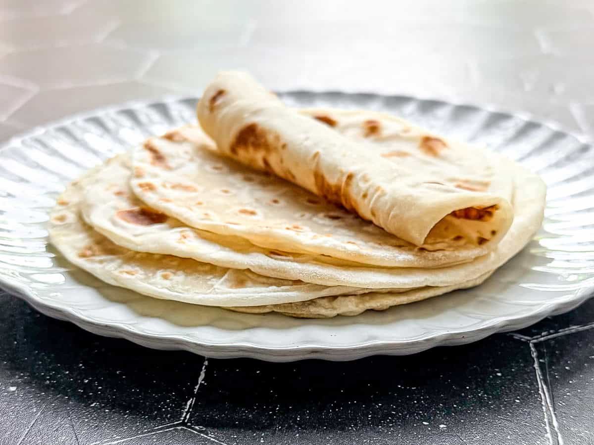 A stack of flour tortillas on a white scalloped edge plate placed on a black tile background.