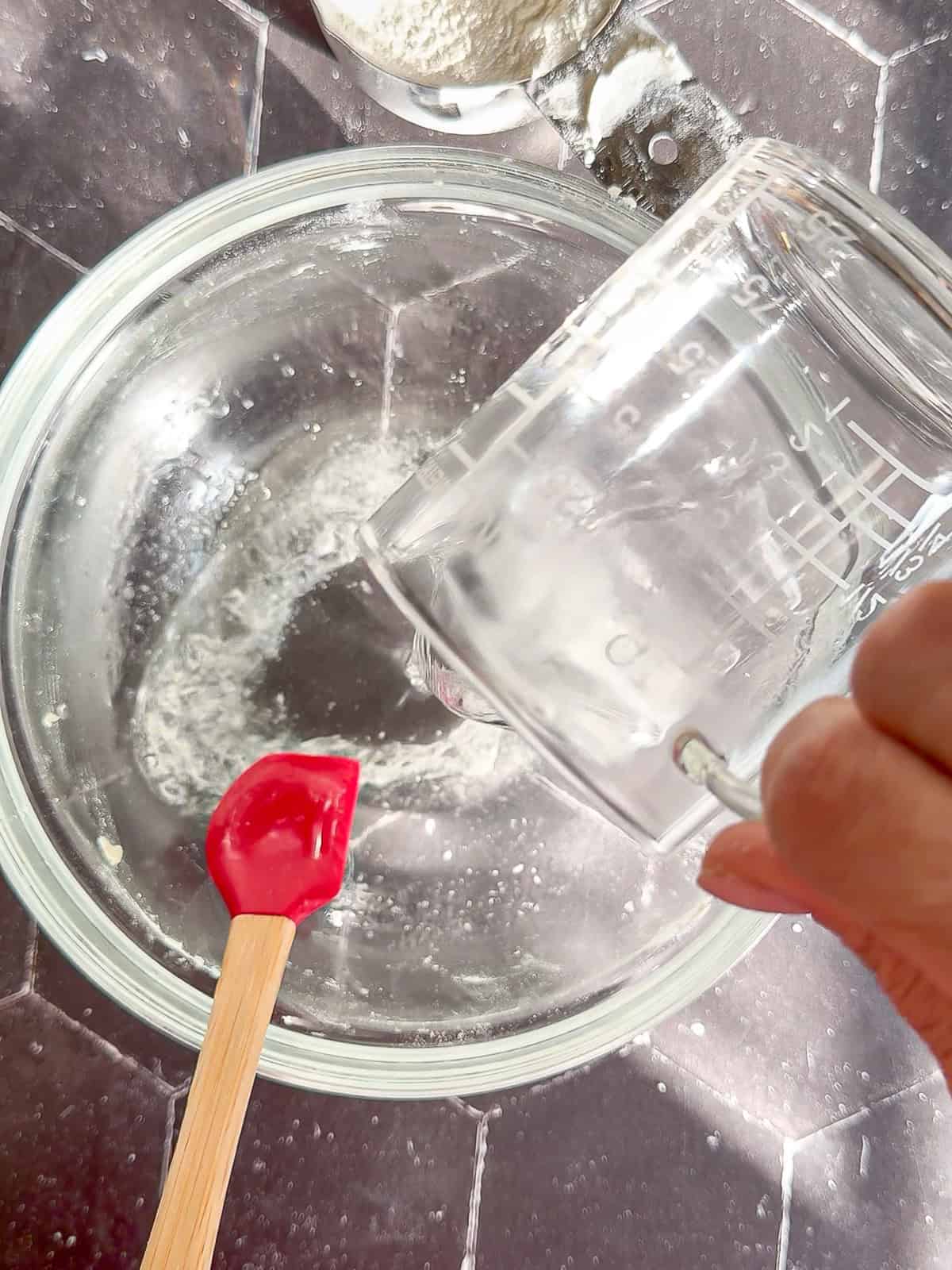 Pouring hot water into melted lard with kosher salt all in a glass bowl on a black tile background.