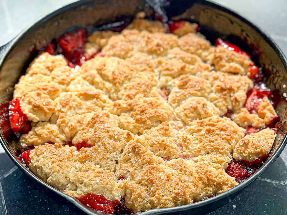 Cast-iron pan containing a strawberry biscuit cobbler on a black tile counter.