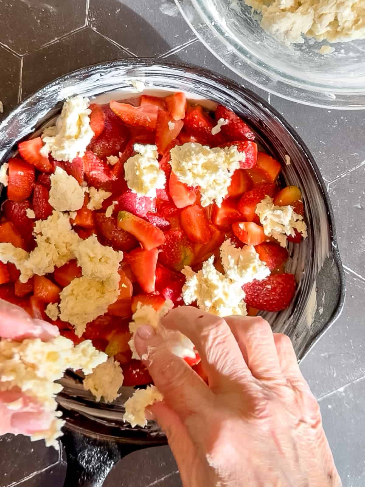 Using fingers to place bits of biscuit dough on top of strawberry filling in a cast-iron pan.