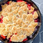 Cast-iron pan containing a strawberry biscuit cobbler on a black tile counter.