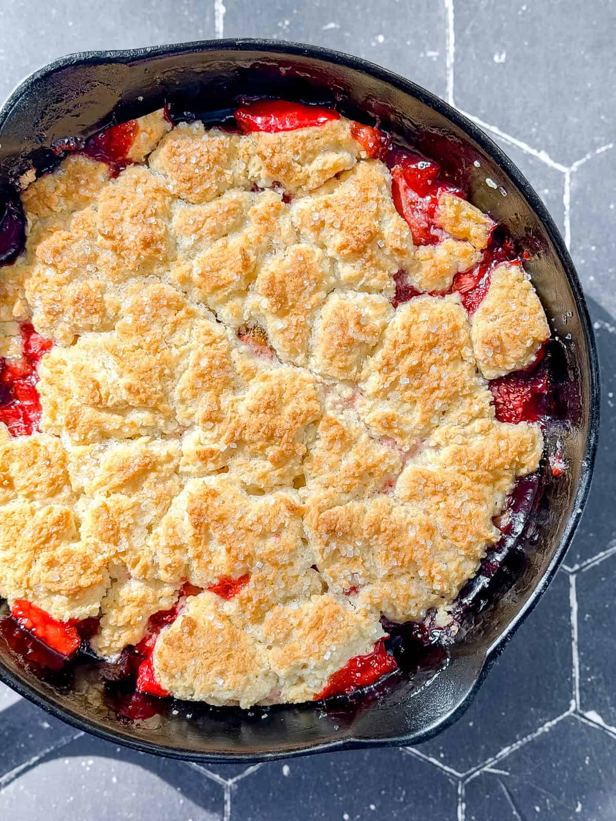 Cast-iron pan containing a strawberry biscuit cobbler on a black tile counter.