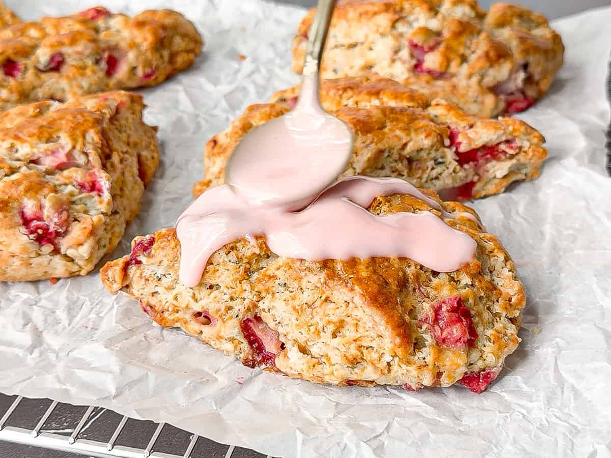 Using a spoon to drizzle a strawberry glaze on top of finished strawberry scones sitting on parchment paper.