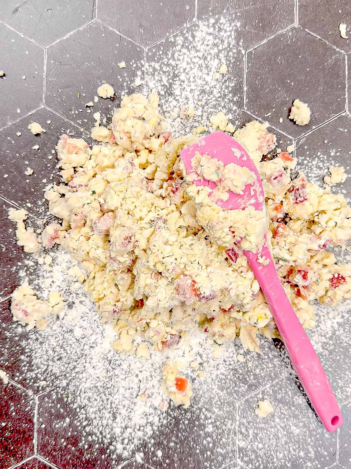 A shaggy and crumbly strawberry scone dough and a pink spatula on a black floured counter.