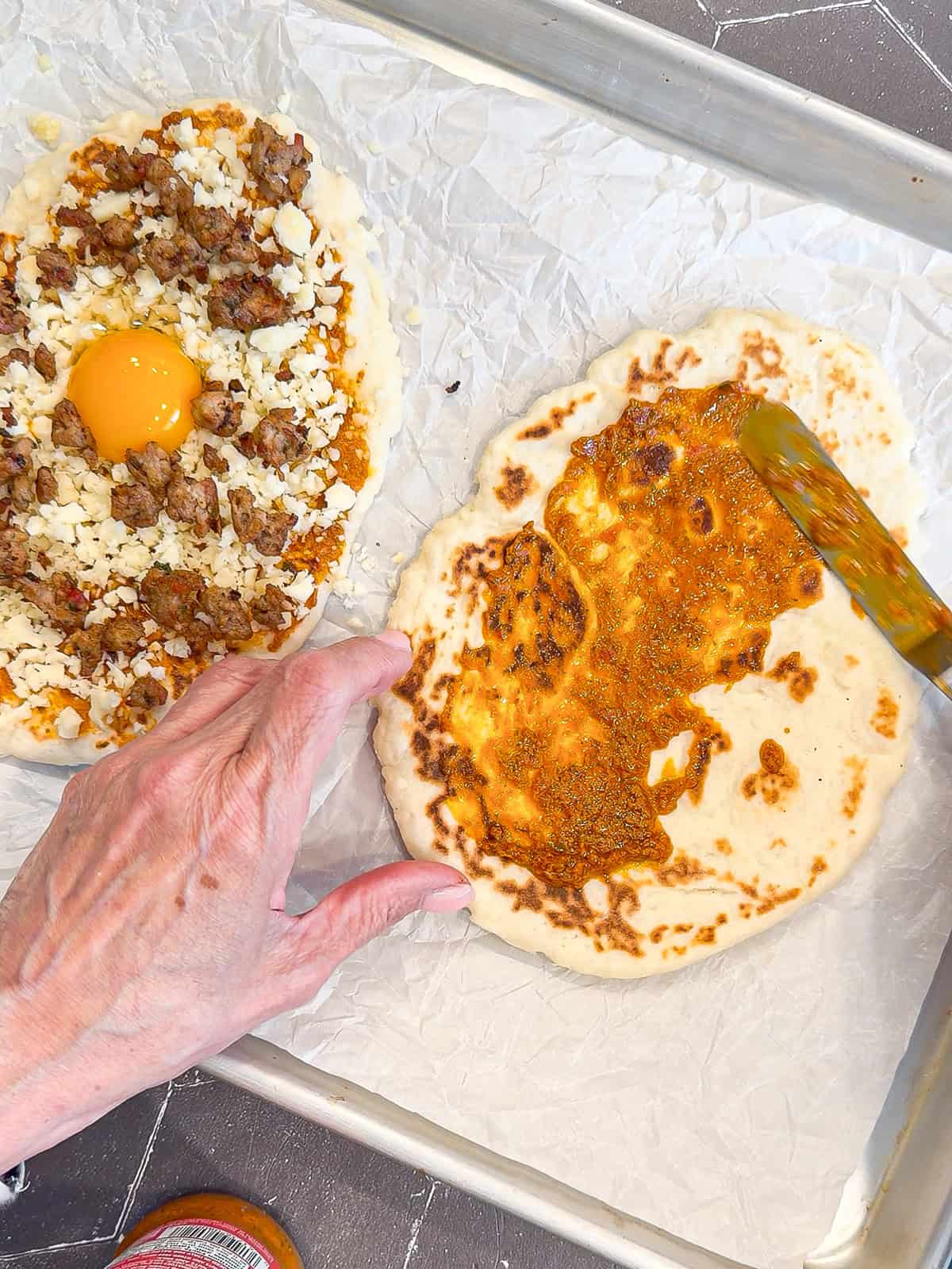 Spreading sun-dried tomato pesto on a piece of flatbread on a parchment paper lined sheet pan.