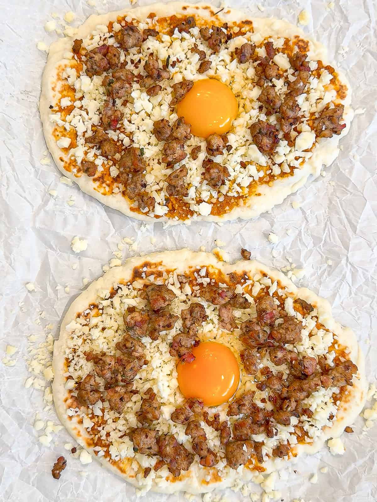 Two flatbread breakfast pizzas on parchment paper ready to be baked.