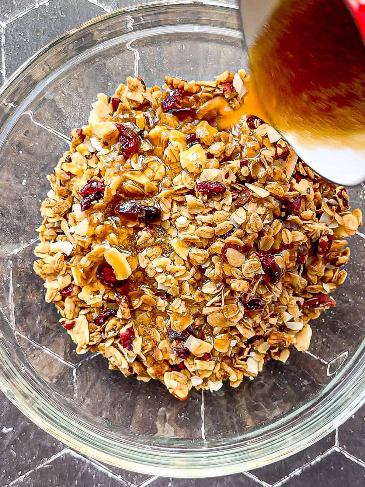 Adding buttery syrup to granola in a glass bowl.
