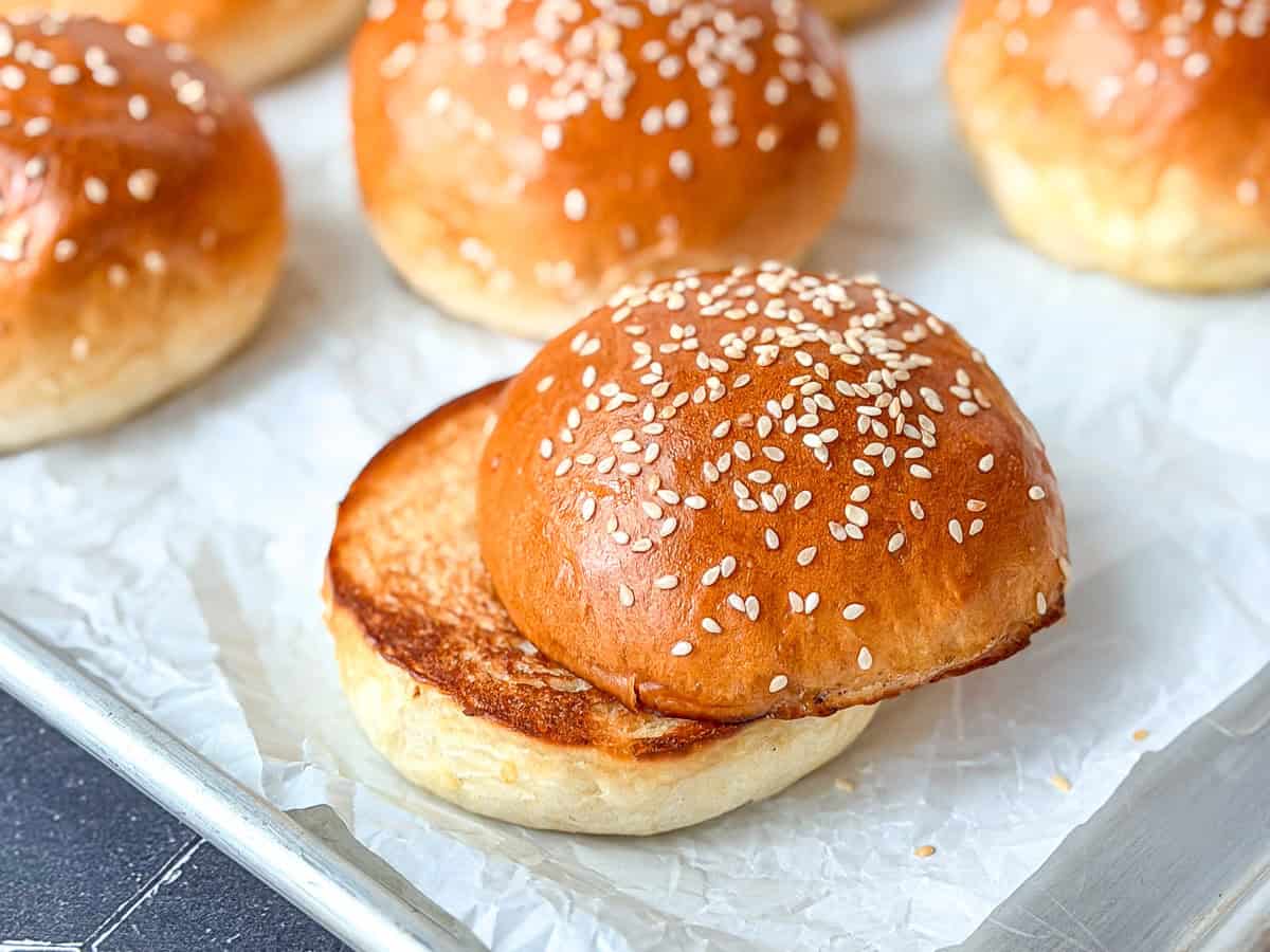 Sesame seed hamburger buns on parchment paper, one of them toasted.