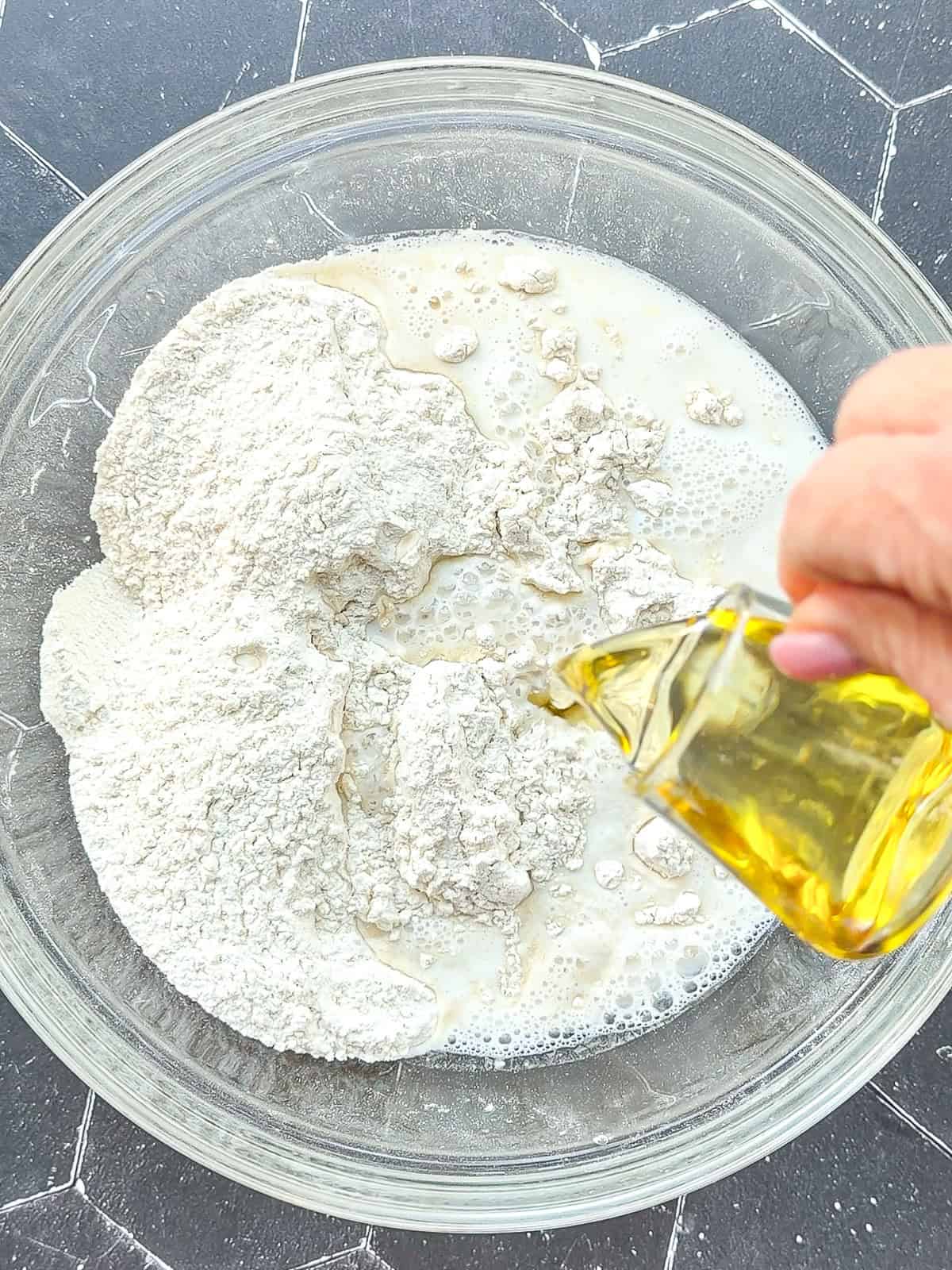 Adding vegetable oil, milk, and water to dry ingredients to make hot dog buns.