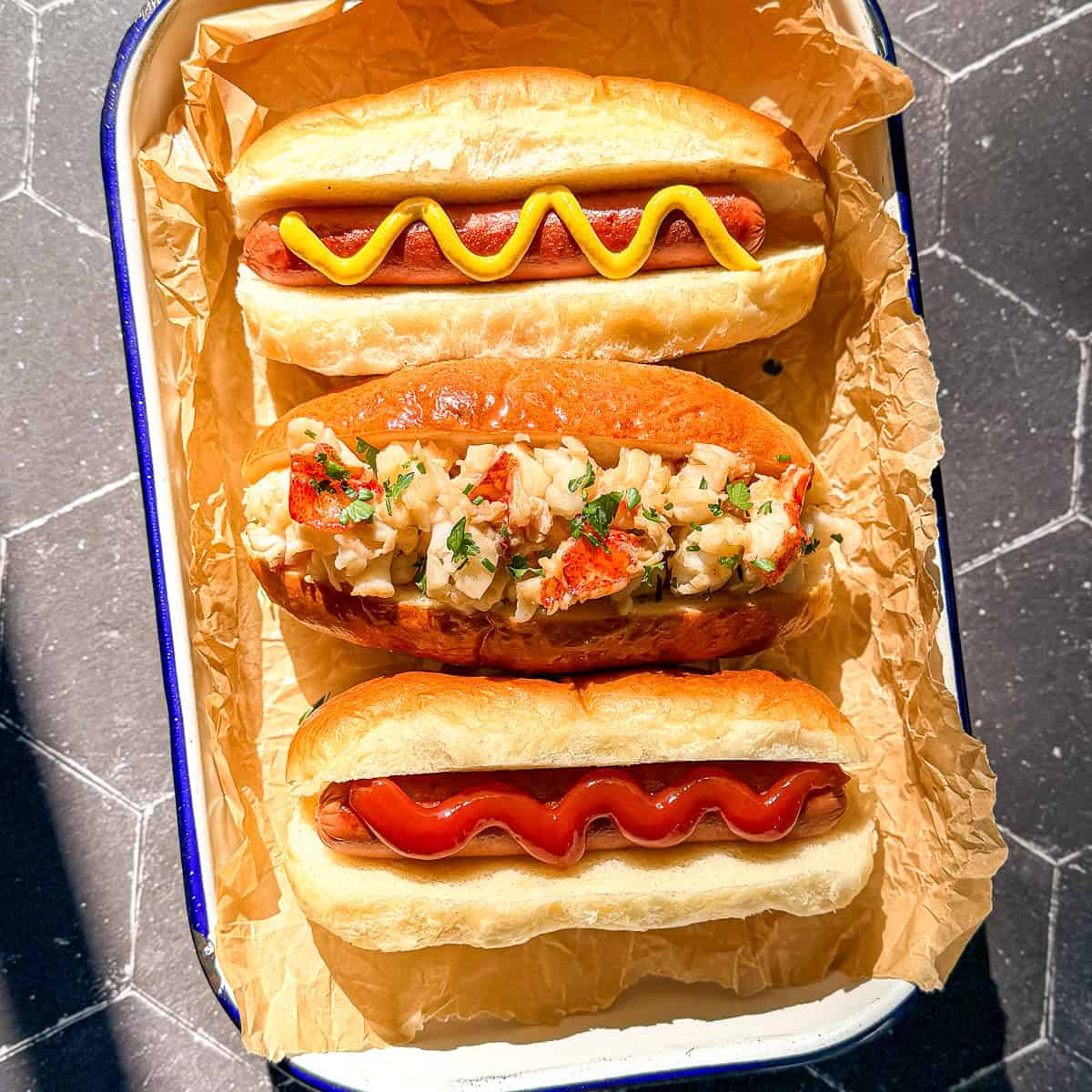 Two hot dogs and a lobster roll on a parchment paper covered pan.