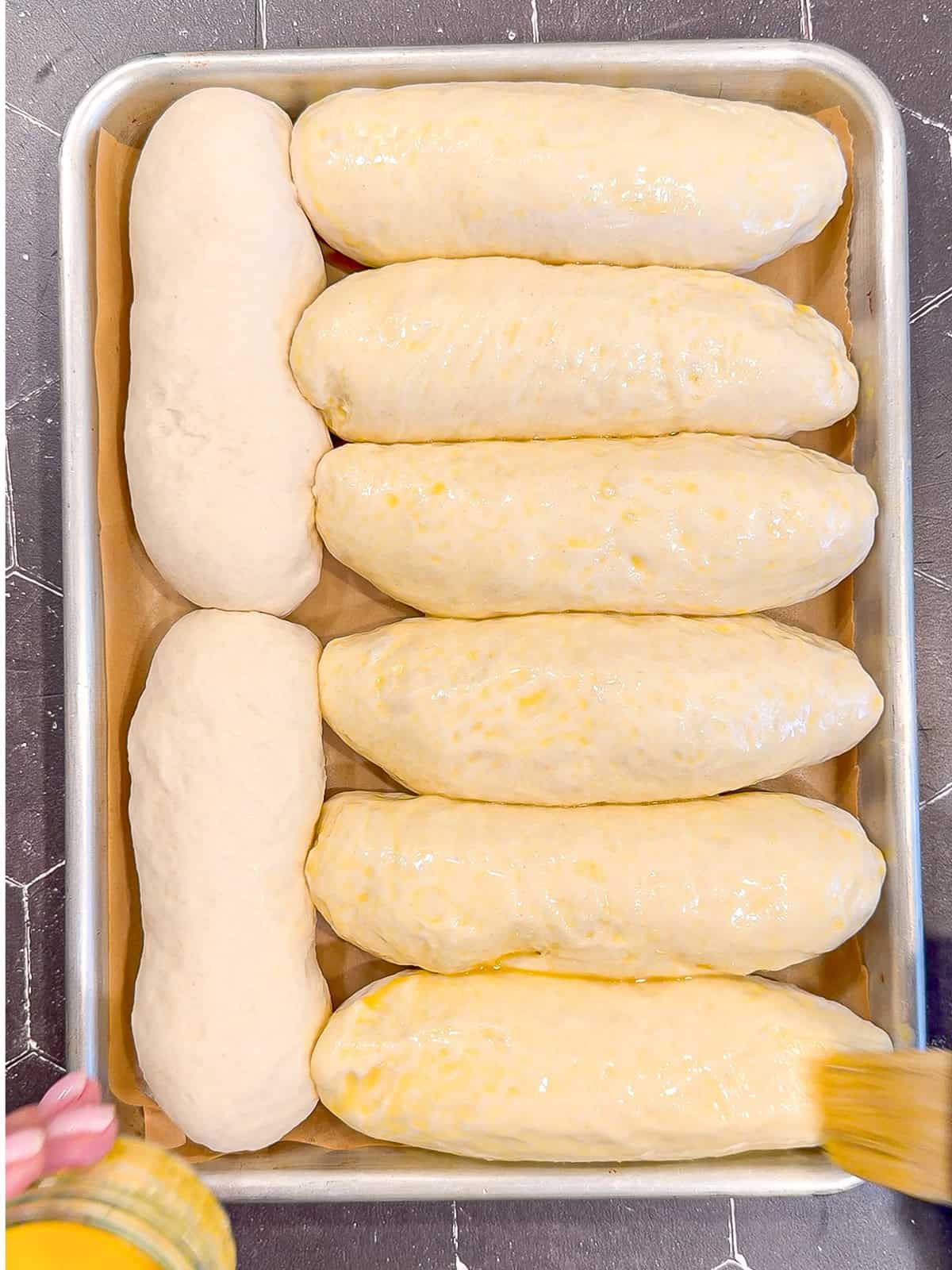 Brushing an egg wash on hot dog buns and a sheet pan.