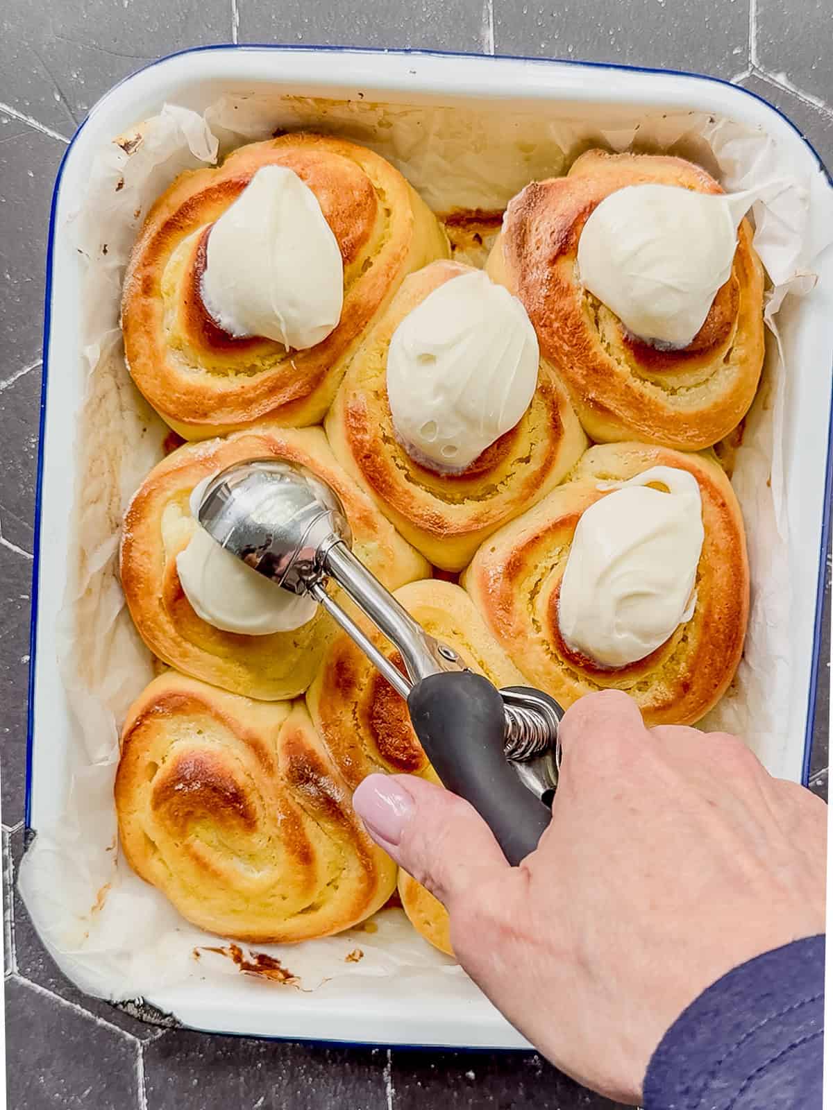 Using a small scoop to Place cream cheese frosting on top of baked lemon rolls in a white pan.
