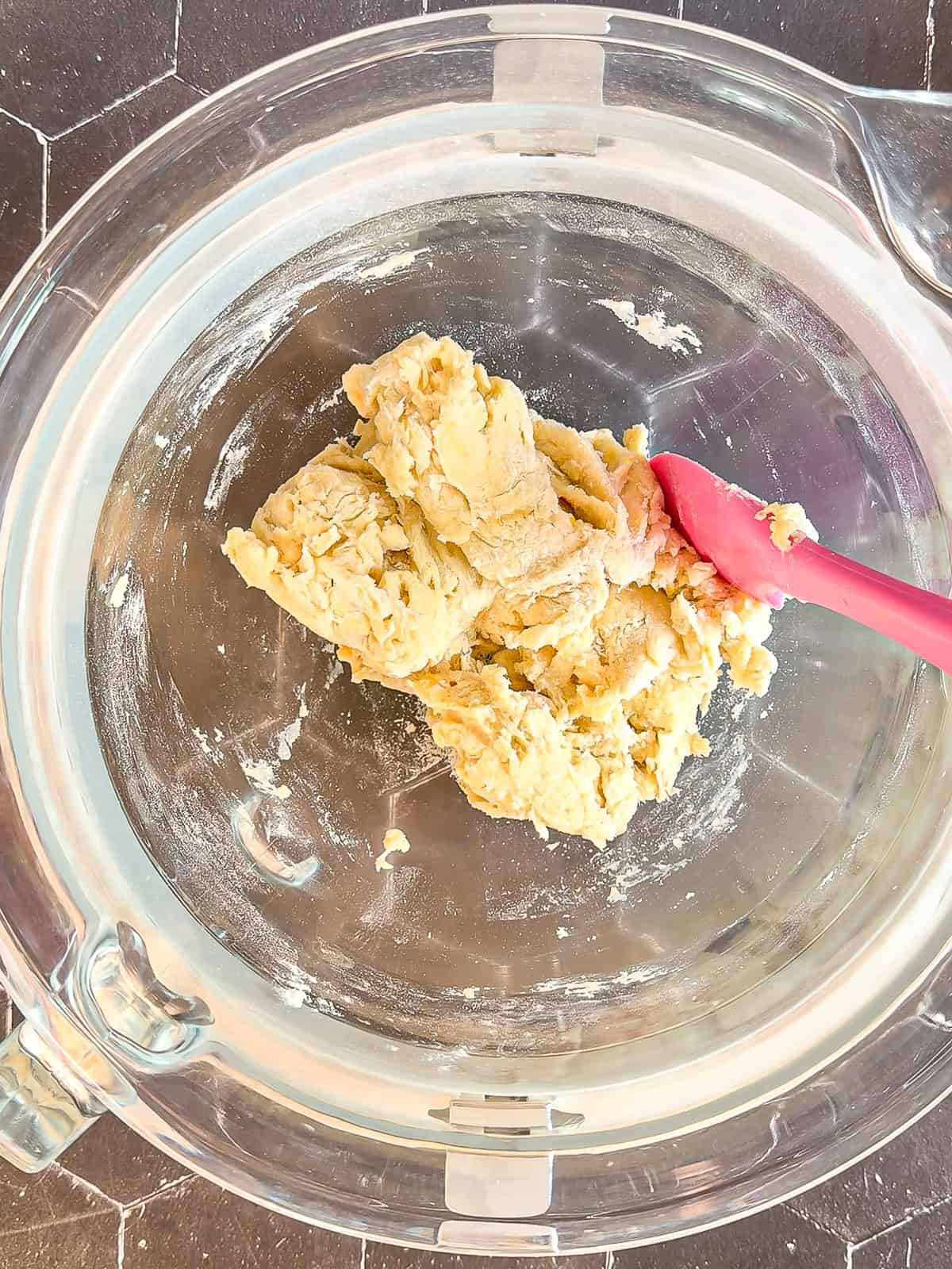 A shaggy mixed dough and a pink spatula in a glass stand mixer bowl.
