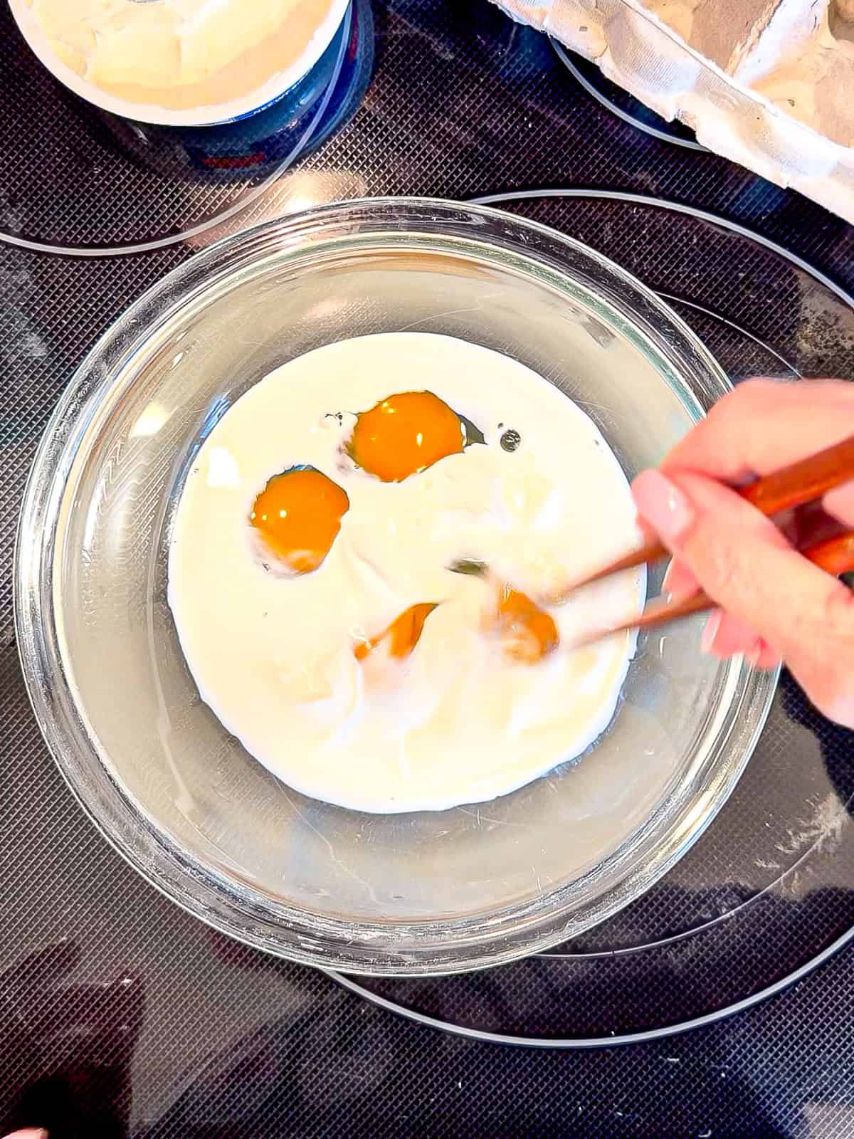 Using chopsticks to whisk eggs, heavy cream, and mascarpone in a glass bowl.