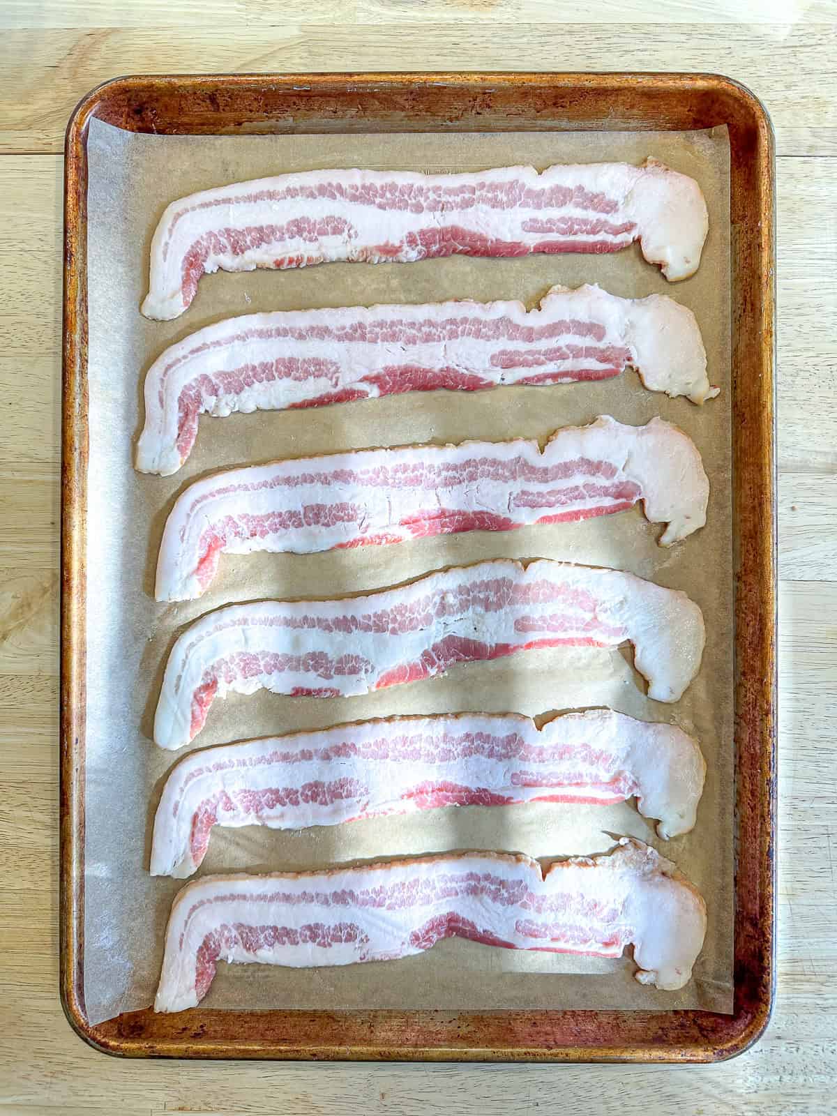 Uncooked bacon strips on a parchment paper lined sheet pan.