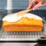 Using a wooden spatula to spread a lemon glaze on top of a loaf cake on a wire rack.
