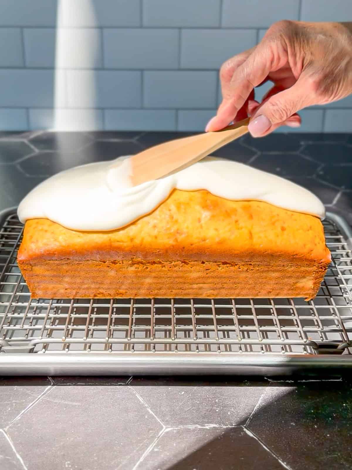 Using a wooden spatula to spread a lemon glaze on top of a loaf cake on a wire rack.
