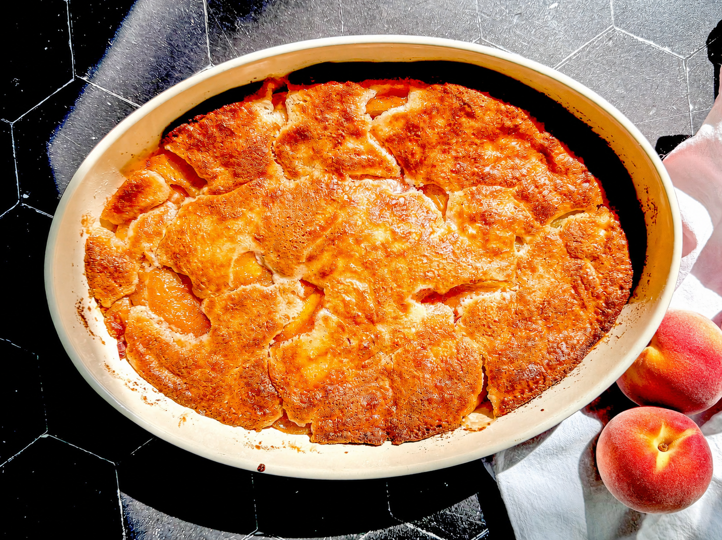 Peach cobbler in an oval dish on a black tile background.