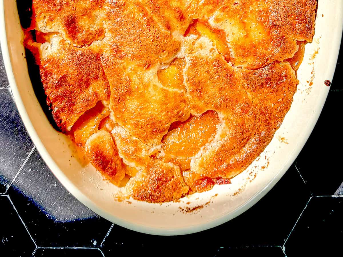 Peach cobbler in an oval dish on a black tile background.
