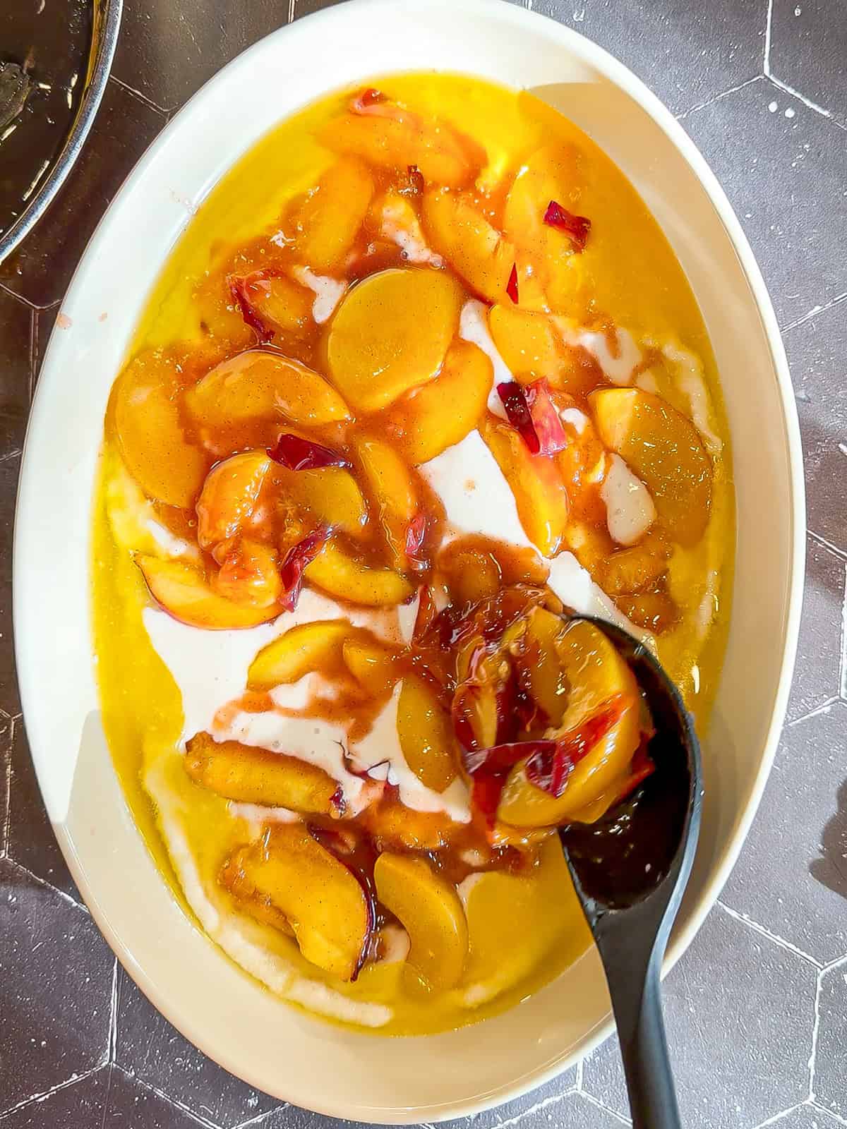 Spooning peach cobbler over batter in an oval baking dish.