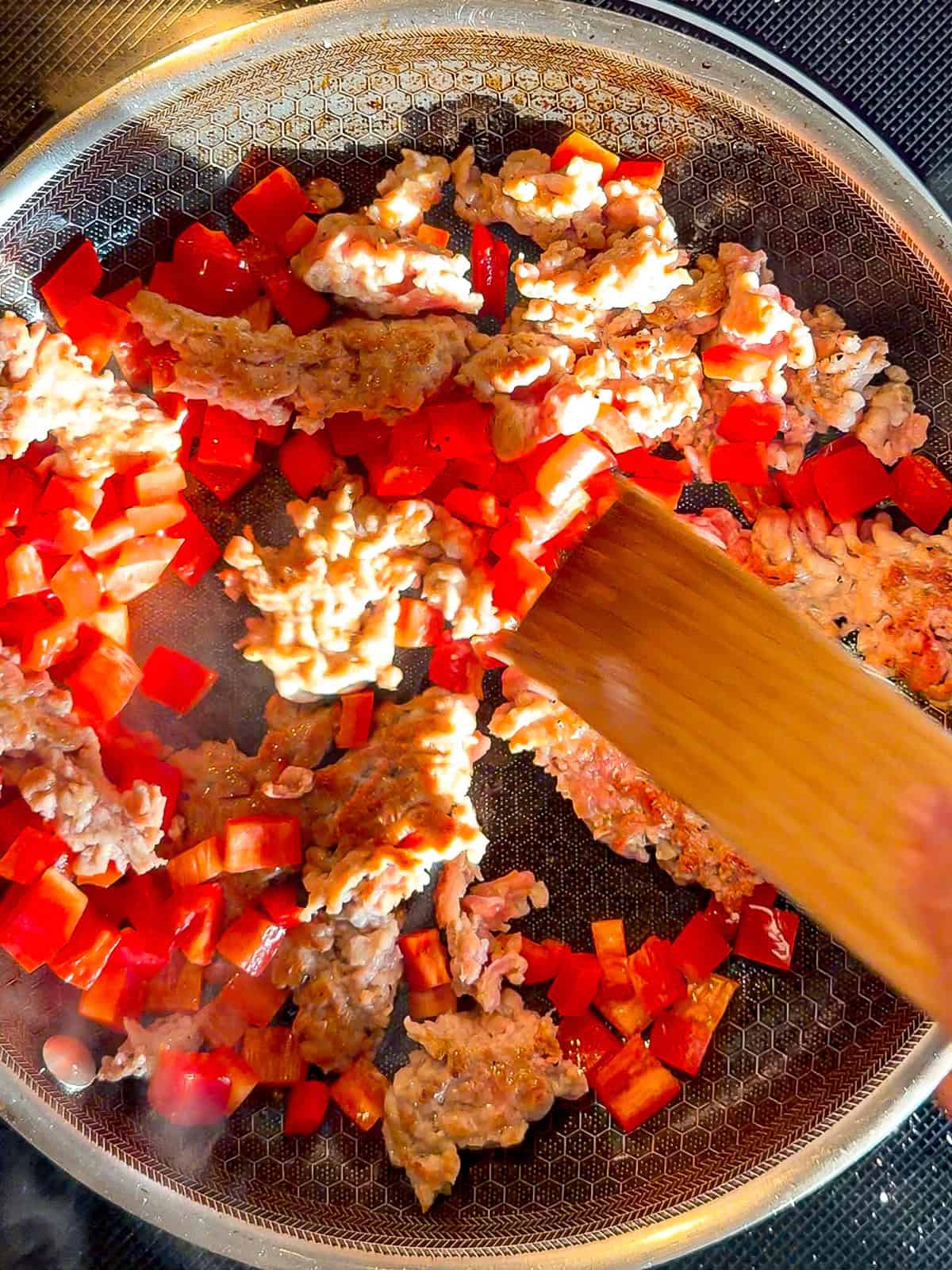 Using a wooden spatula to stir diced bell pepper and Italian sausage in a black pan.