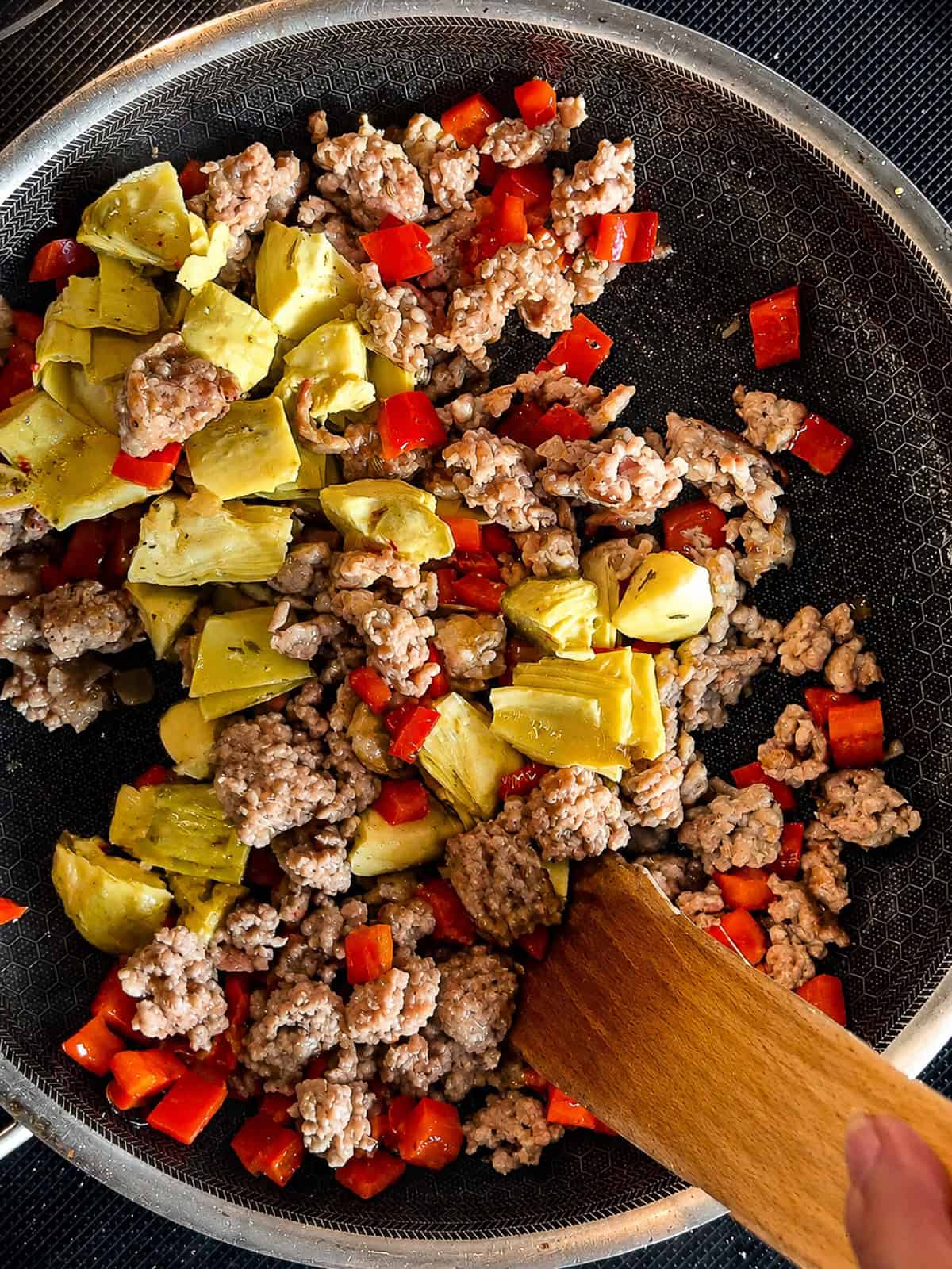 Sausage, red peppers, and artichoke hearts in a pan cooking.