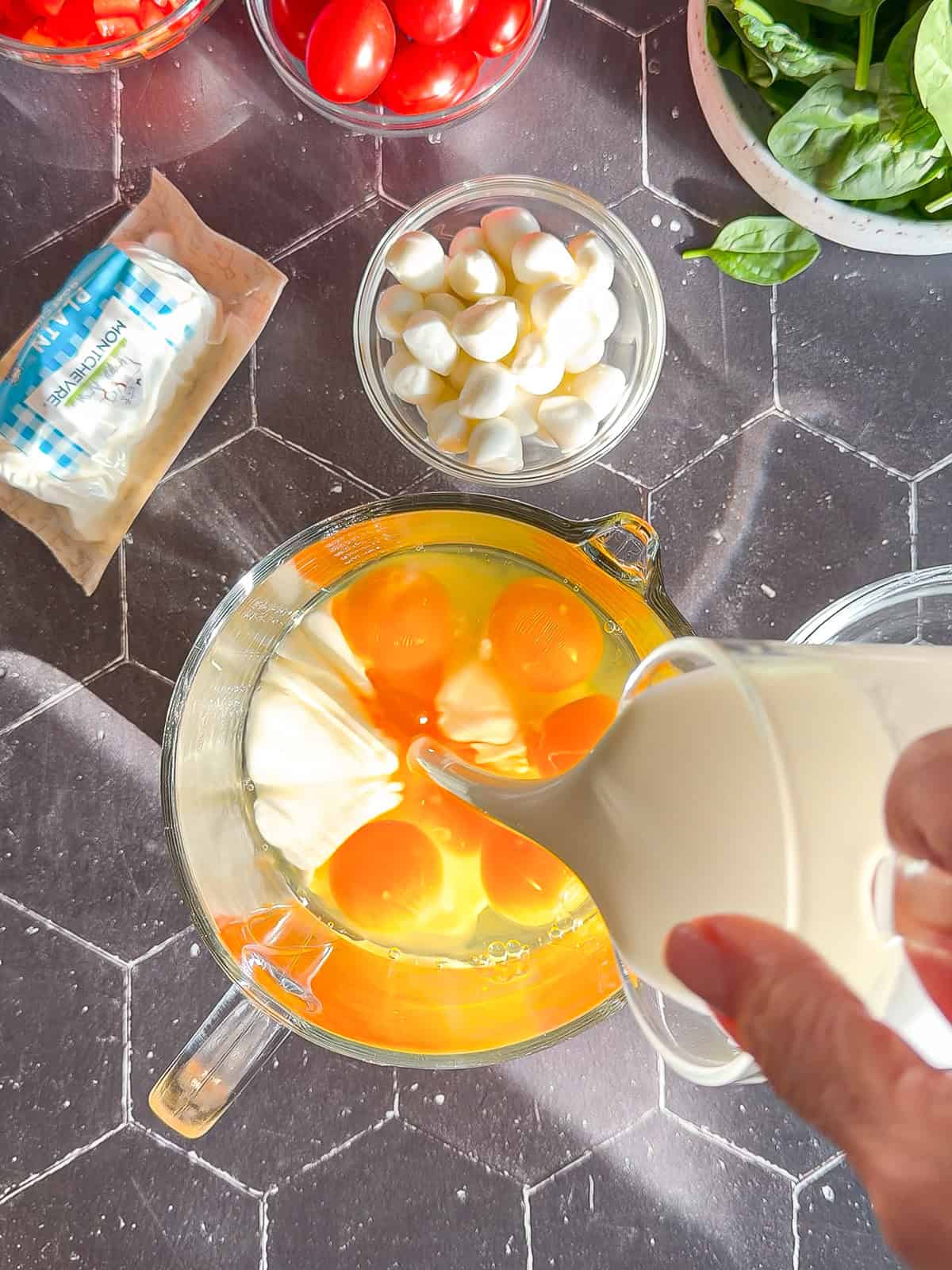 Pouring heavy cream into a jug filled with eggs, with cheese and assorted vegetables next to them on a black counter.