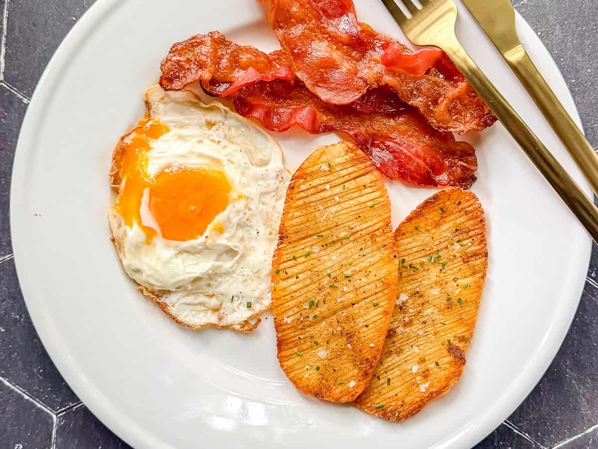 Two crispy accordion potatoes on a white plate with bacon and eggs.