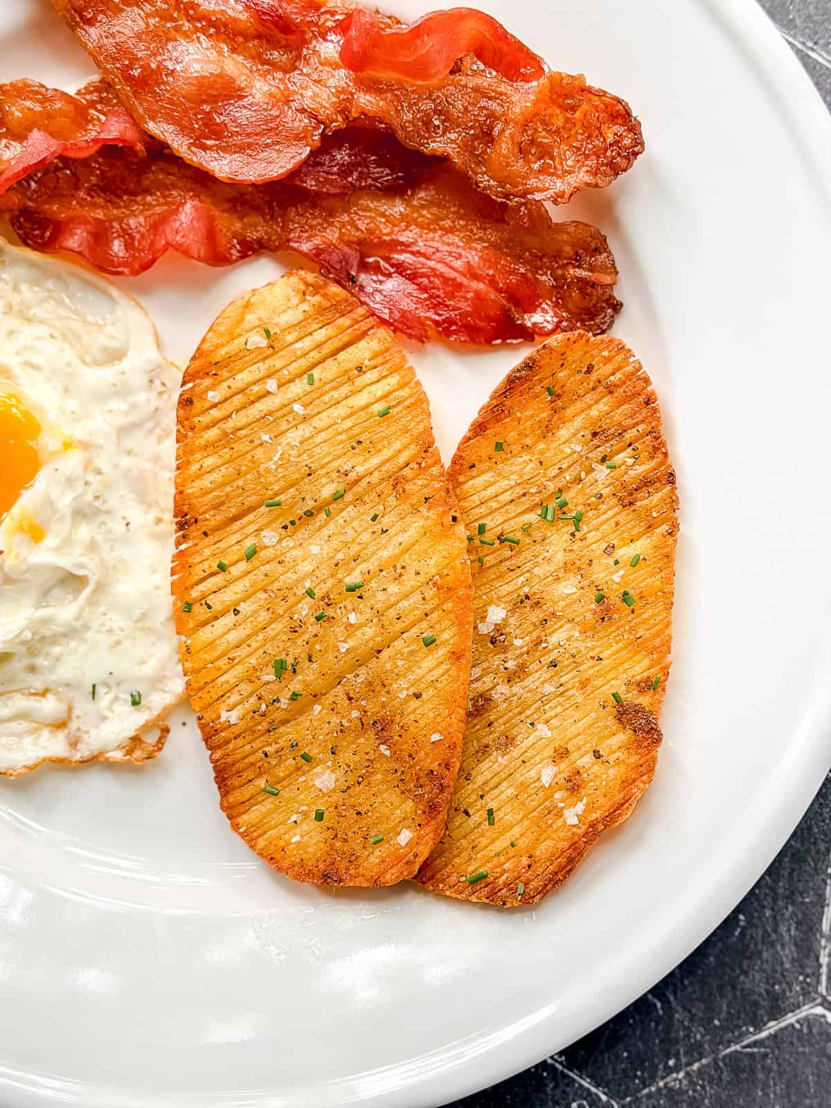 Two crispy accordion potatoes on a white plate with bacon and eggs.