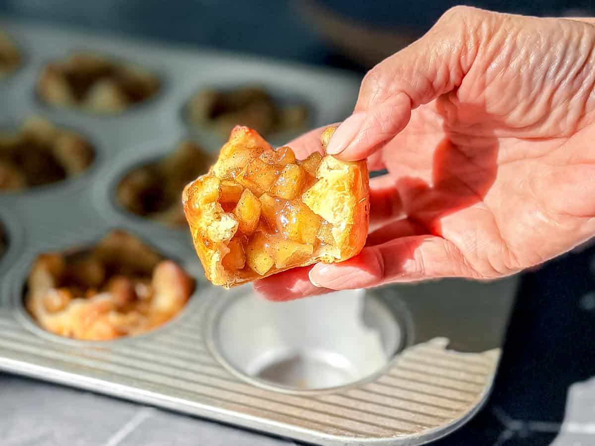 Hand holding a baked apple croissant over a muffin pan filled with the same.