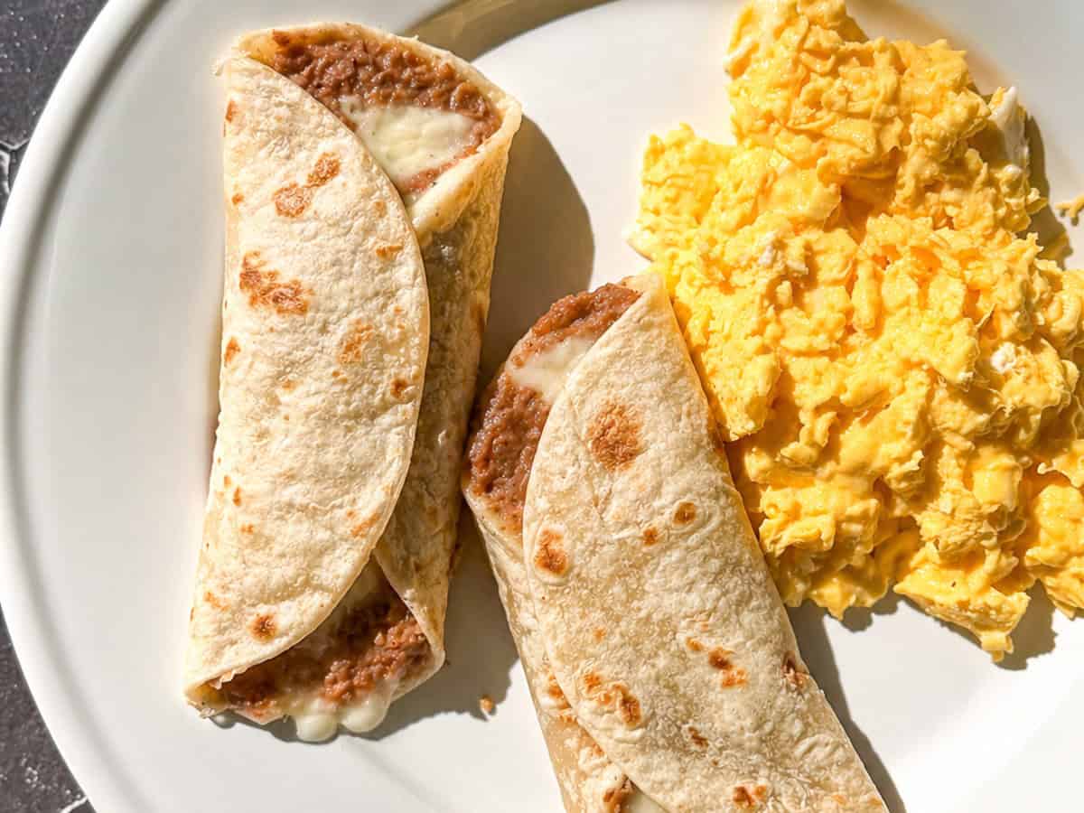 Two bean and cheese tacos next to scrambled eggs on a white plate.