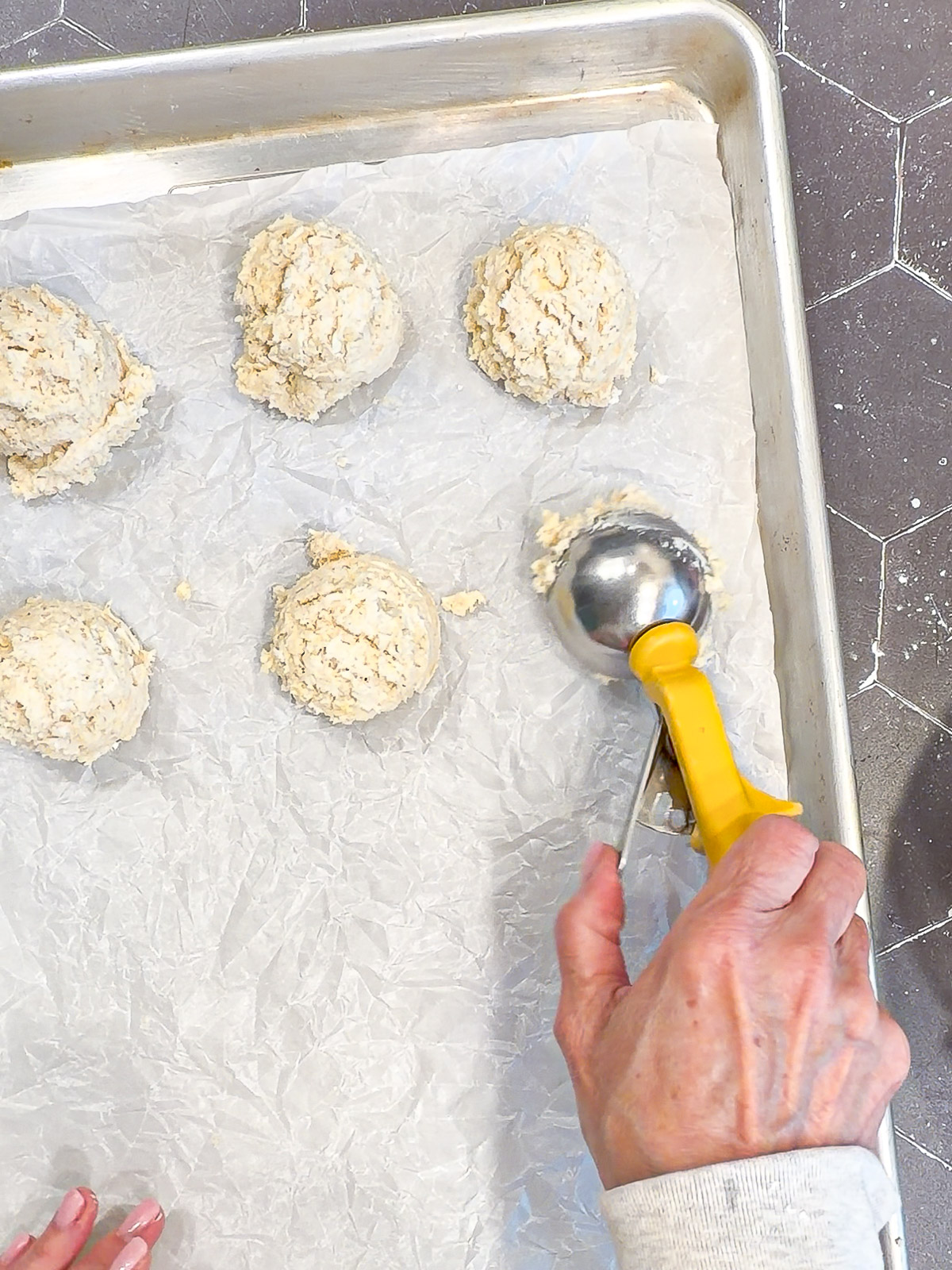 Using an ice cream scoop to portion biscuit dough onto a parchment paper lined sheet pan.