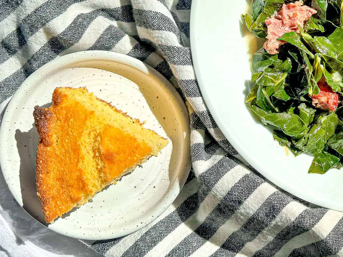 A slice of cornbread next to a bowl of collard greens on a blue and white striped napkin.