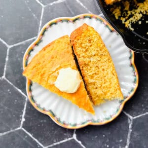 Two slice of cornbread on a scalloped plate next to a pan of cornbread.