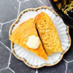 Two slice of cornbread on a scalloped plate next to a pan of cornbread.