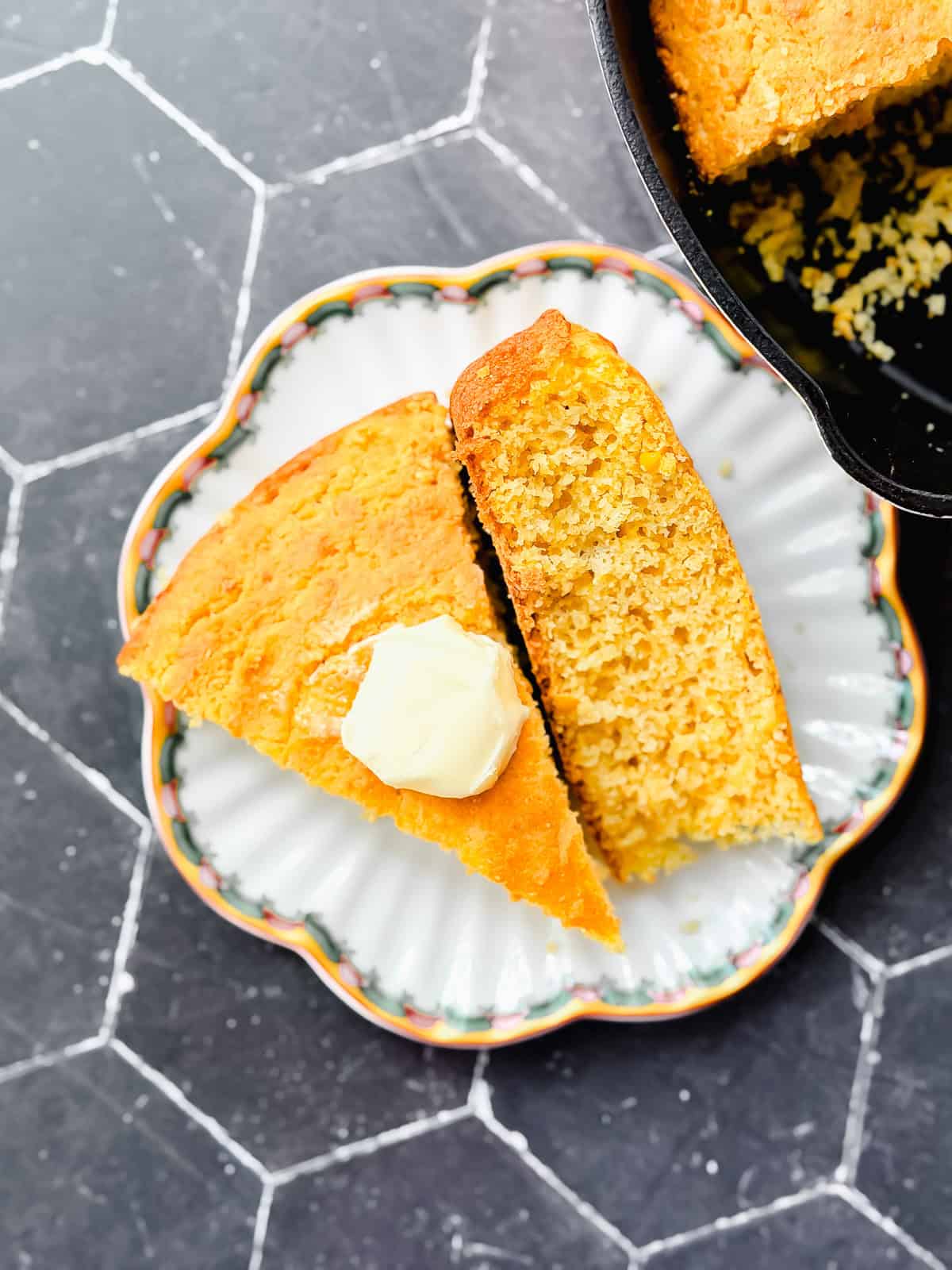 Two slice of cornbread on a scalloped plate next to a pan of cornbread.