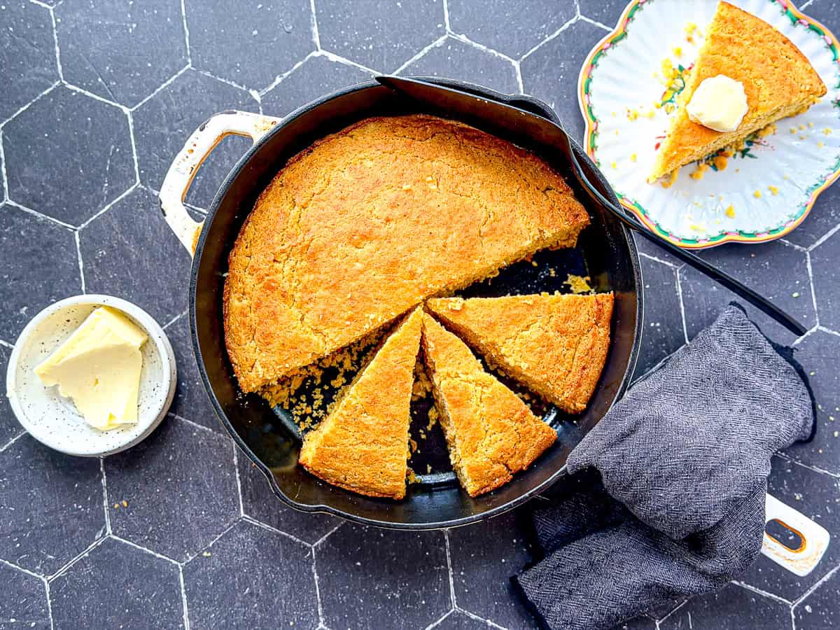 A skillet of sliced cornbread on a black tile background.