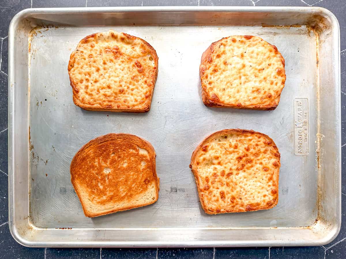 Four slices of cheese toast on a sheet pan, one turned over to show the crispy bottom.