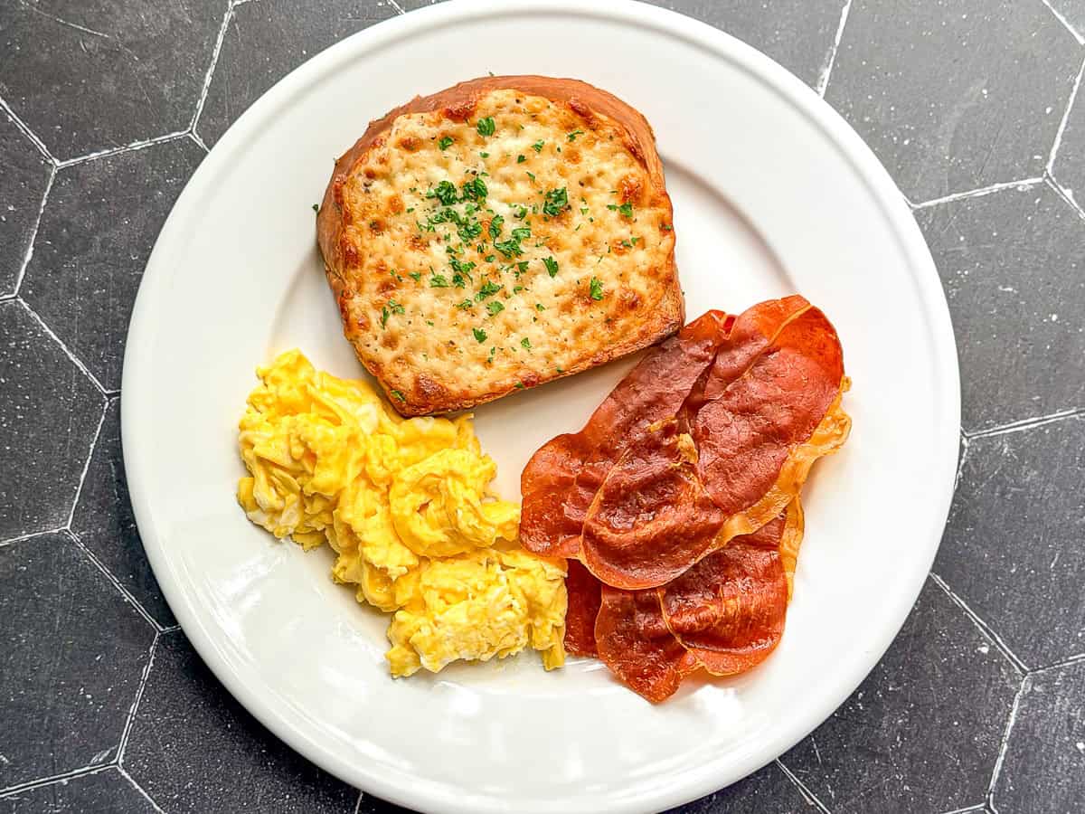 Cheese toast with scrambled eggs and crispy prosciutto on a white plate.